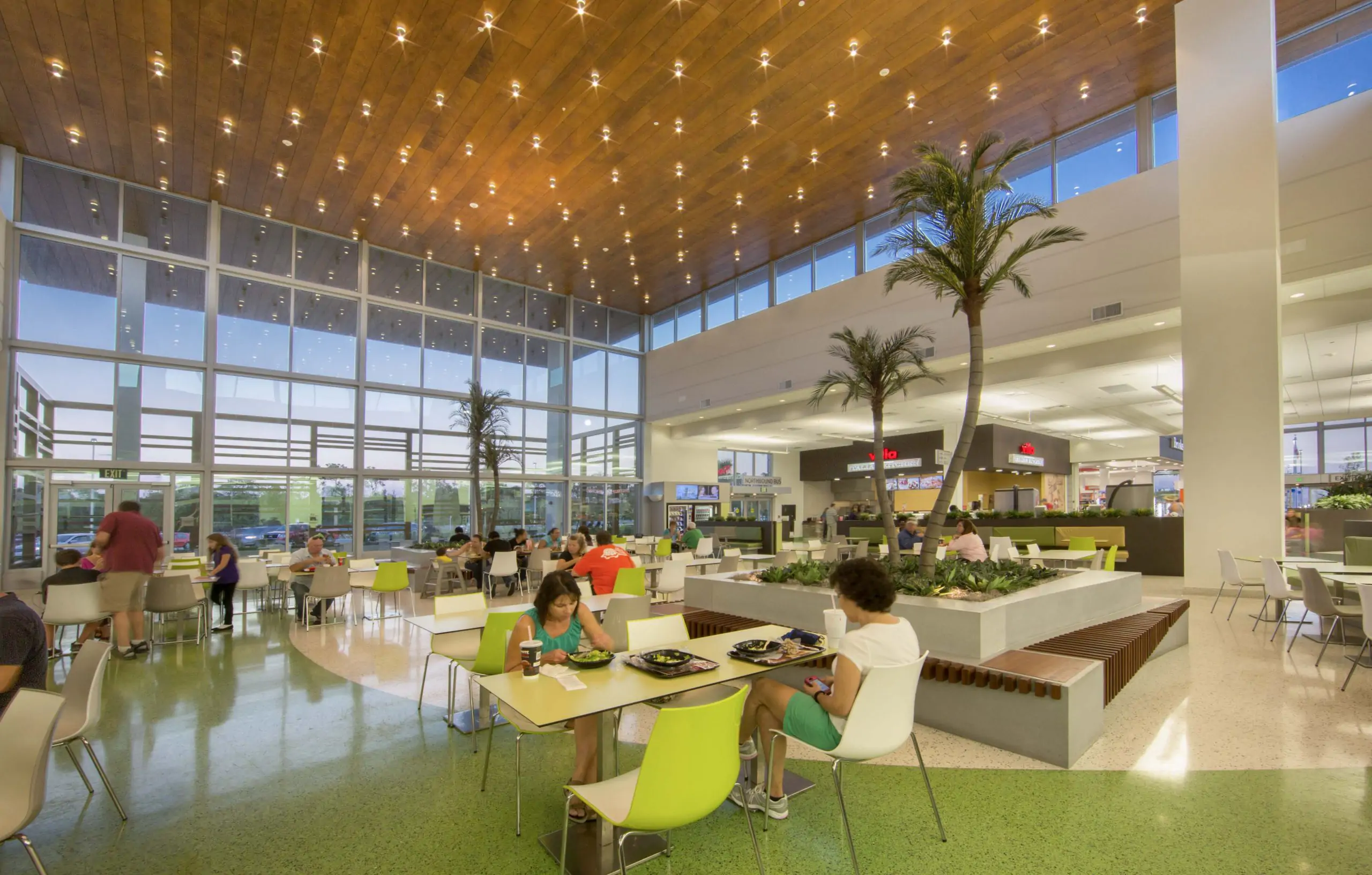 Modern airport food court with high wooden ceiling, palm trees, and people dining at tables.