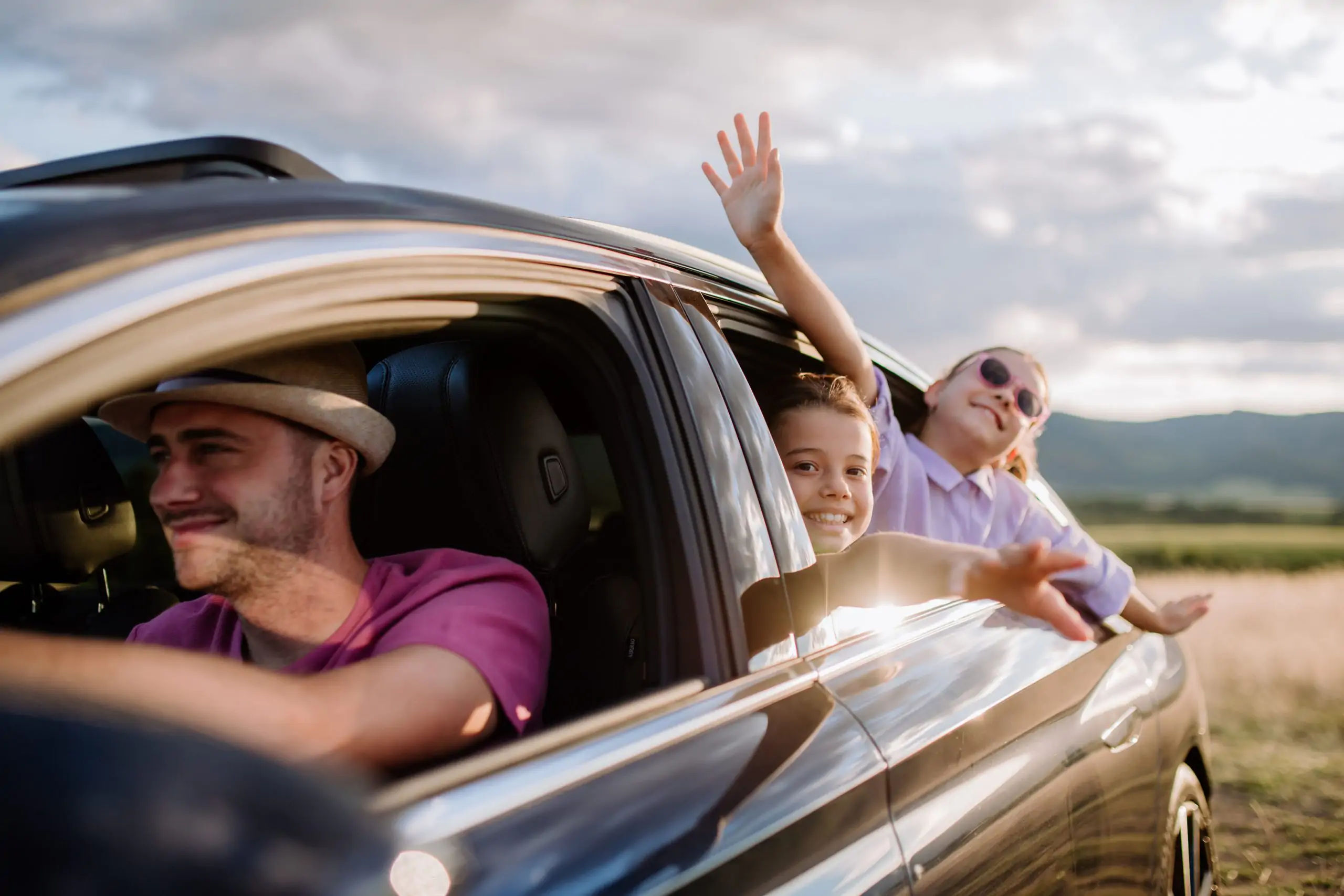 Family enjoying a road trip, children waving from the car window at sunset.