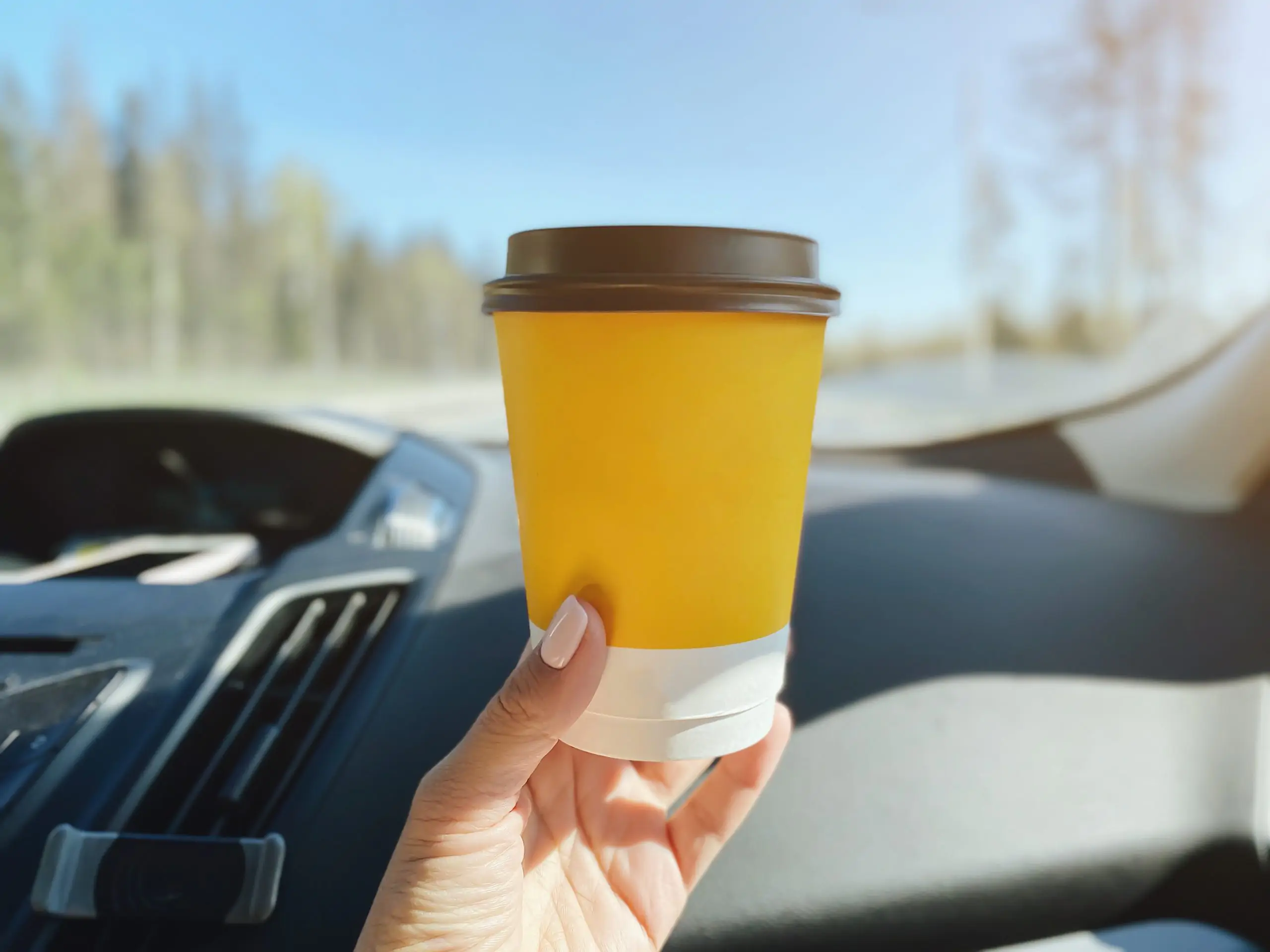 Hand holding a yellow takeaway coffee cup inside a car on a sunny day.