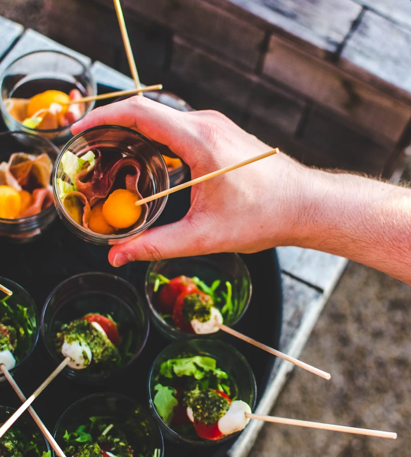 Hand holding a cup of appetizers with skewers at a casual dining setting.